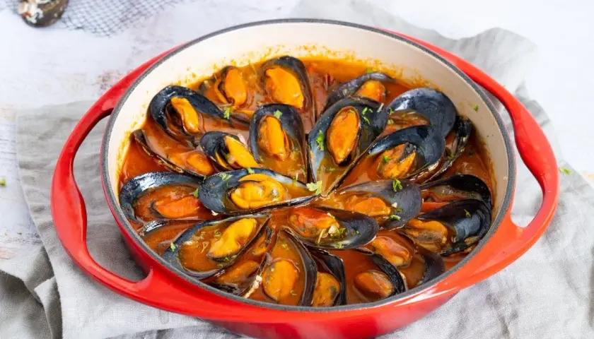 Mejillones en salsa roja, servidos en una cazuela roja, con perejil picado espolvoreado.