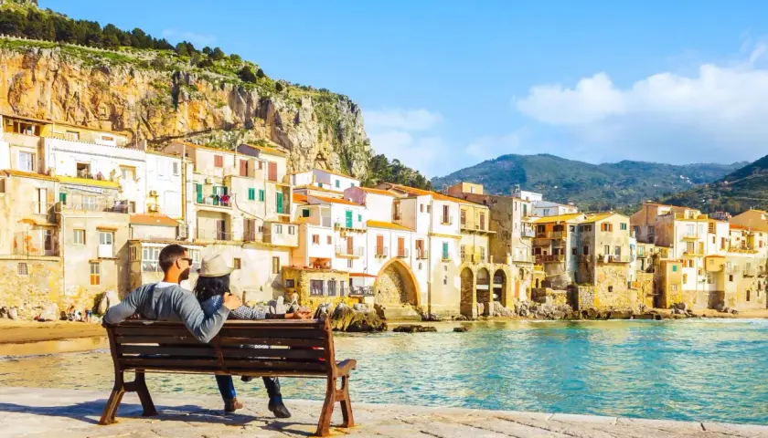 Coppia seduta su una panchina, ammira il mare e le case colorate di Cefalù, un luogo perfetto per vacanze in Sicilia sul mare.