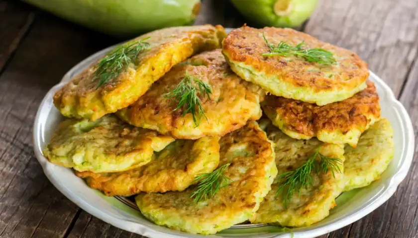 Pile de beignets de courgette dorés et moelleux, garnis de brins d'aneth frais, sur une assiette blanche. Deux courgettes vertes floues en arrière-plan.