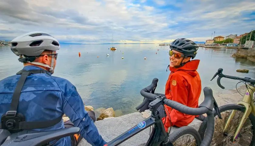 Two cyclists enjoy a break during bike guided tours in the Mediterranean, with boats on the calm sea and a coastal town in the background.