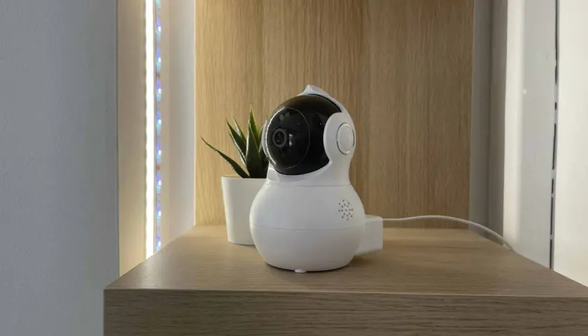 Cámara de vigilancia blanca con lente negra, parte de un sistema domótico, junto a una planta y luces LED.