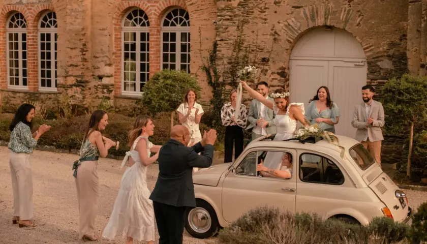 Mariage Dolce Vita : la mariée, radieuse, lève son bouquet depuis une Fiat 500. Ses invités applaudissent devant un manoir en pierre.