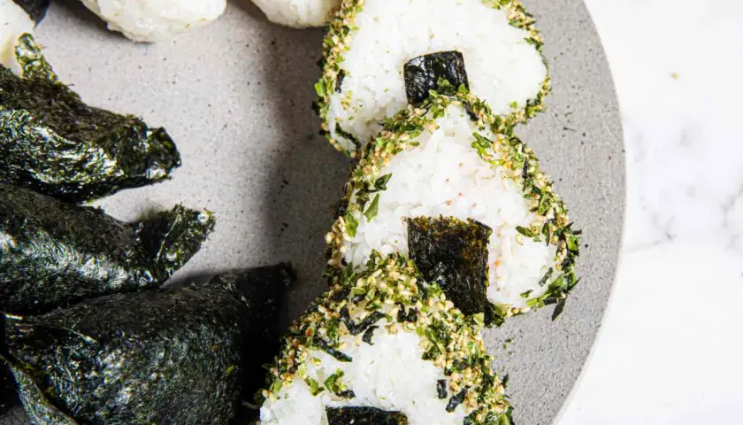 Triangular sushi rice balls, some wrapped in nori, others coated in sesame seeds and herbs, are artfully arranged on a grey plate.