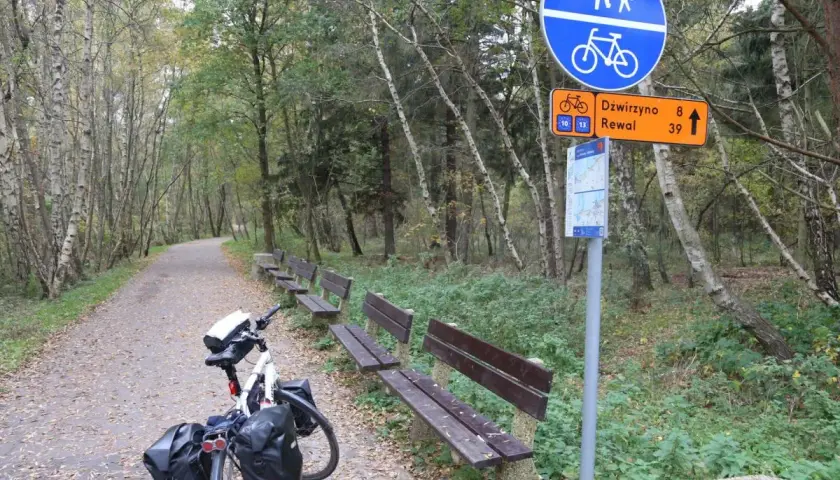 Ścieżka rowerowa Velo Baltica w lesie, z ławkami i znakiem wskazującym Dźwirzyno i Rewal. Długość trasy zachęca do podróży.