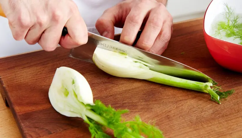 Hände schneiden Fenchel für ein Gericht mit feinem Fenchel Geschmack. Ein Teil liegt auf dem Holzbrett, ein anderer im roten Schälchen.