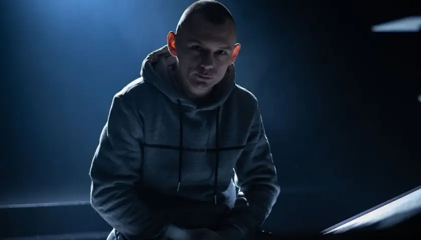 Młody polski raper w szarej bluzie z kapturem, oświetlony od tyłu niebieskim światłem.