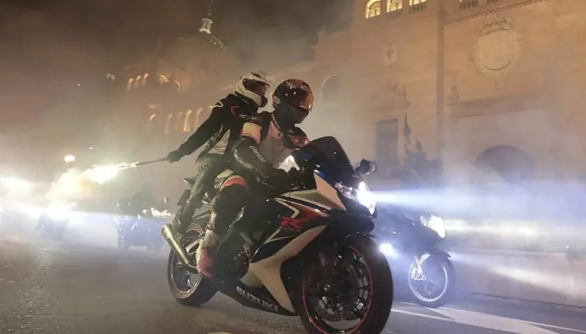 Dos motoristas en una Suzuki, uno con una bengala encendida, circulan por la noche. Parece que una motocicleta puede circular sin tubo de escape, creando un espectáculo de luces y humo.