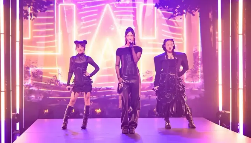 Trzy artystki k-pop w czarnych, stylizowanych strojach na scenie z neonowym tłem.