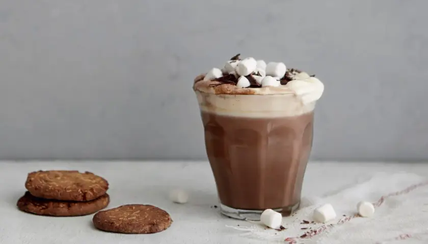 En härlig varm choklad med fluffigt skum, marshmallows och chokladströssel. Bredvid ligger några krispiga kakor.