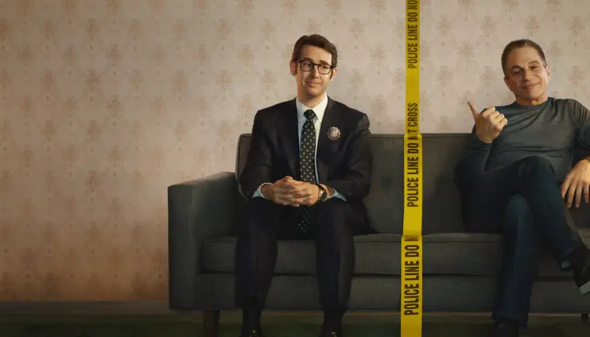 Dwóch mężczyzn siedzi na kanapie. Jeden, ubrany w garnitur, to **the good cop**. Obok niego, za taśmą policyjną, drugi mężczyzna uśmiecha się.