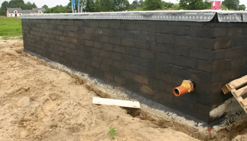 Pomarańczowa rura kanalizacyjna przechodzi przez fundament z czarnej cegły, przygotowywany do dalszych prac budowlanych.
