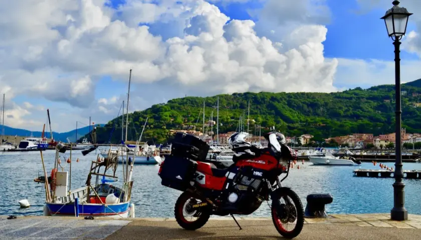Moto rossa pronta a partire dal porto azzurro dell'Elba, con barche a vela e colline verdi sullo sfondo.