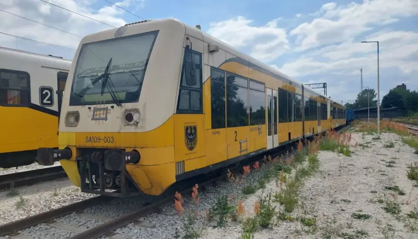 Żółty pociąg SA109-003 stoi na torach, gotowy do podróży.