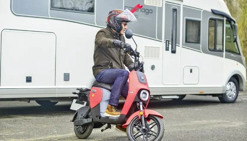 Un homme casqué conduit un scooter rouge devant un camping-car. Ce scooter est parfait pour explorer les environs lors de vos voyages en camping-car.