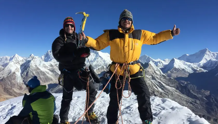 Dwóch alpinistów na szczycie Mera Peak, jeden z czekanem, drugi z kciukiem w górę. W tle ośnieżone góry.