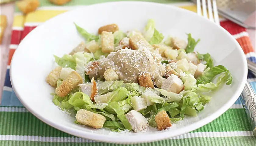 Una deliciosa ensalada cesar con pollo, crutones crujientes y queso parmesano rallado, servida en un plato blanco sobre un mantel colorido.