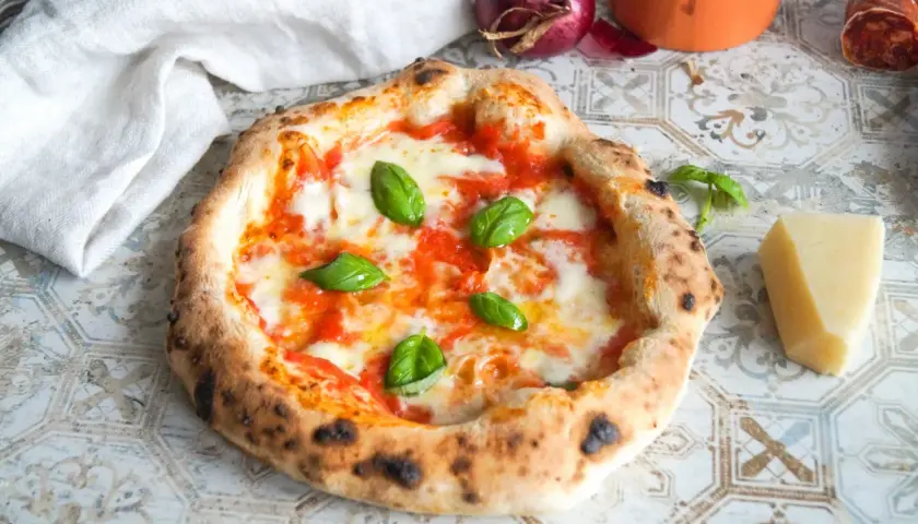 Eine authentische Pizza Napoletana mit frischem Basilikum, geschmolzenem Mozzarella und Tomatensauce. Daneben ein Stück Parmesan und eine rote Zwiebel.