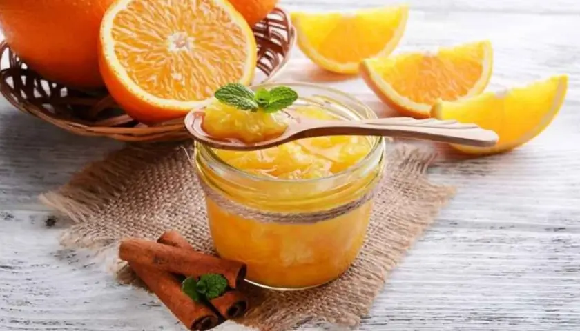 Tarro de mermelada de naranja casera, con naranjas frescas y canela en rama. Una cucharada muestra la textura.