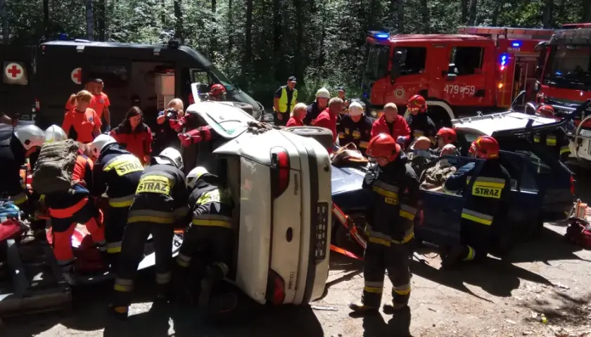 Akcja ratunkowa ksrg po wypadku. Strażacy wyciągają poszkodowanych z przewróconego samochodu. W tle wozy strażackie i karetka.