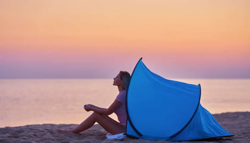 Kobieta siedzi na plaży przy rozłożonym namiocie plażowym. Zachód słońca nad morzem. Jak złożyć namiot plażowy instrukcja.