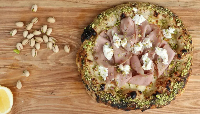 Pizza mit Mortadella, Pistazien und cremigem Käse, daneben einige Pistazien und eine Zitronenspalte.