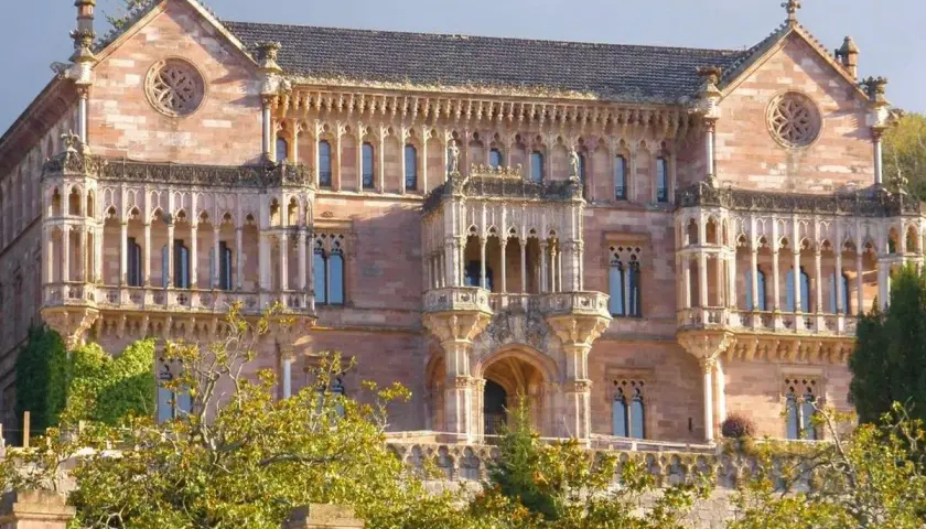 El **Palacio de Comillas**, una joya arquitectónica de piedra rosada, se alza majestuoso entre la vegetación, con sus balcones y rosetones detallados.