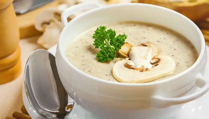Aksamitna zupa krem z pieczarek, udekorowana plasterkiem pieczarki i gałązką natki pietruszki, czeka na łyżkę.