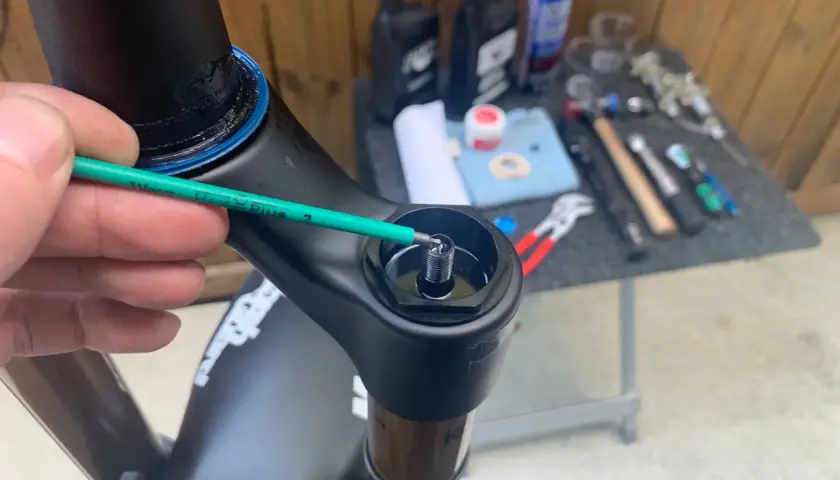 A hand uses a green hex wrench to adjust a bolt on a black bicycle fork, part of a Fox 34 service. Tools and lubricants are visible in the background.