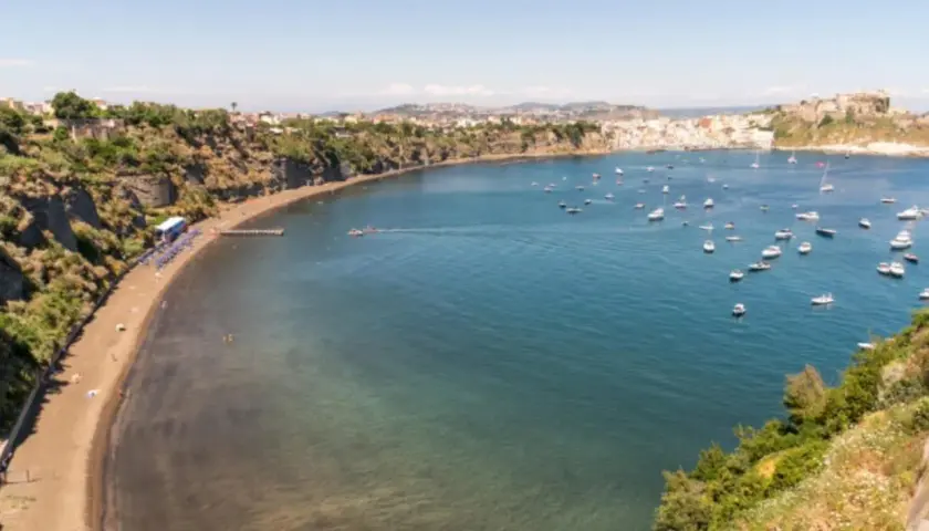 Vue d'une baie pittoresque, avec une plage de sable noir, des falaises verdoyantes et une multitude de bateaux dans l'eau bleue. Un paysage qui rappelle la voisine de Capri.