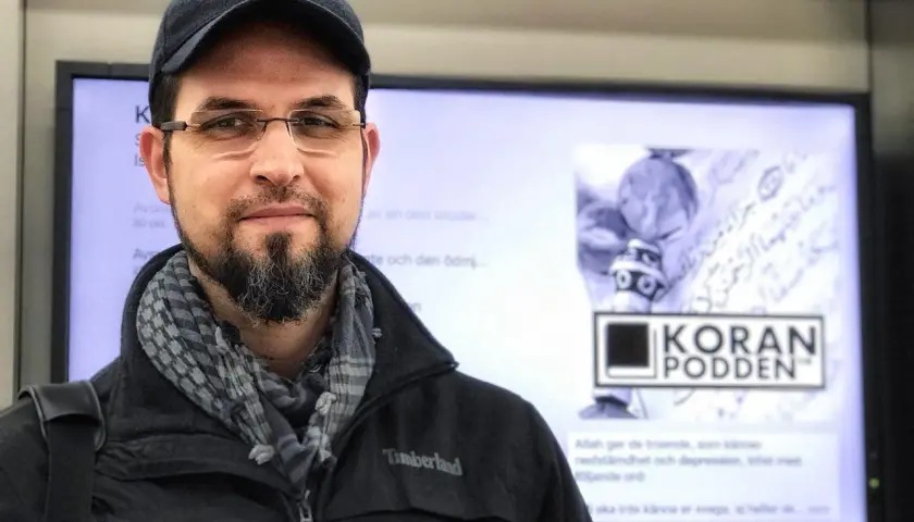 Man med skägg och keps ler mot kameran. I bakgrunden syns en skärm med texten "KORAN PODDEN" och arabisk skrift. Allahu alam.