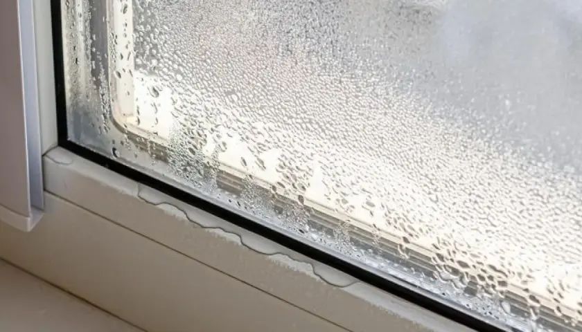 Gotas de agua cubren el cristal de la ventana, indicando condensación. El alféizar muestra pequeñas acumulaciones de agua.