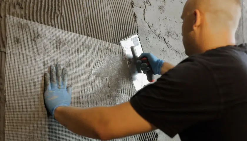 Hand mit blauer Handschuh drückt Gittergewebe in Mörtel auf der Wand. Dies hilft gegen Feuchtigkeit in der Wand.