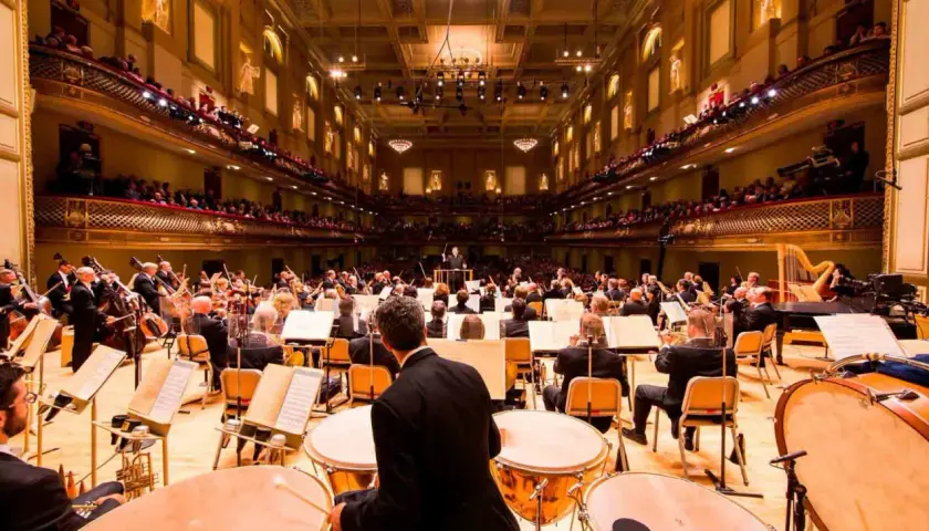 Un orchestre symphonique interprète de la musique classique moderne dans une salle de concert ornée, avec des musiciens jouant de divers instruments.