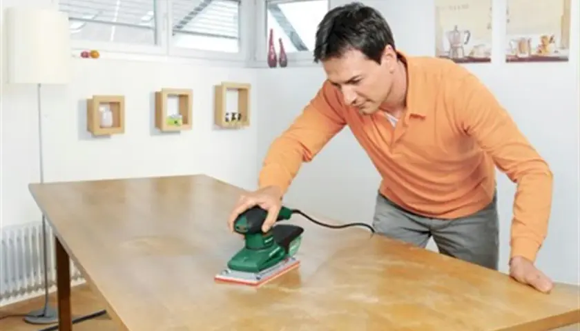 Un homme utilise une ponceuse vibrante ou excentrique pour rénover une table en bois.