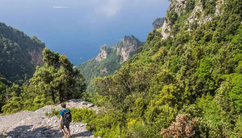 A hiker walks along the rocky path of the Sentiero degli Dei, with lush green mountains and the blue sea stretching out below.