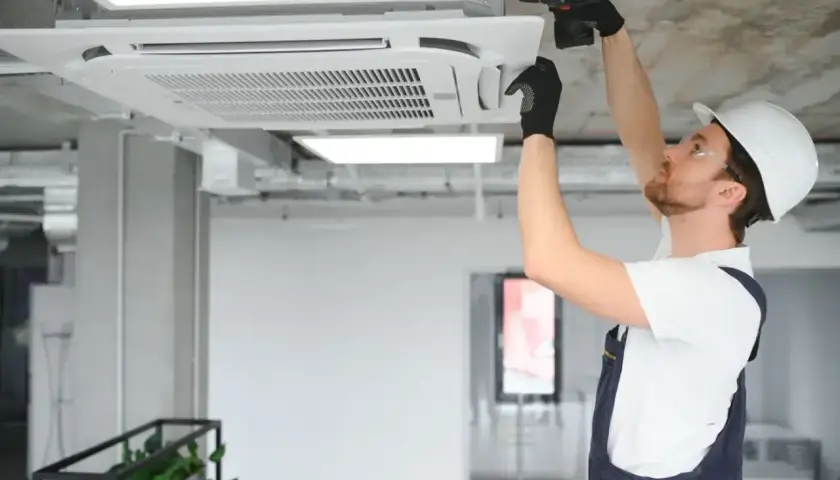 Instalator w kasku montuje klimatyzator kasetonowy na suficie.