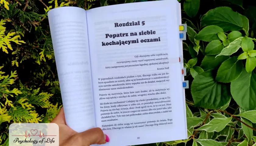 Otwarta książka "Popatrz na siebie kochającymi oczami" w otoczeniu zieleni. Tekst mówi o walce z negatywną samokrytyką i brakiem akceptacji siebie.