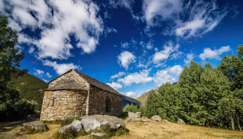 Pequeña ermita de piedra en las rutas del Vall de Boí, bajo un cielo azul con nubes algodonosas.