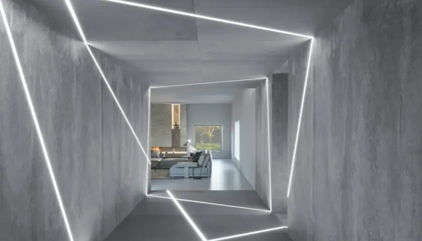 Nowoczesny korytarz z betonowymi ścianami i geometrycznym podświetleniem ściany od góry. W tle salon z kominkiem i kanapą.