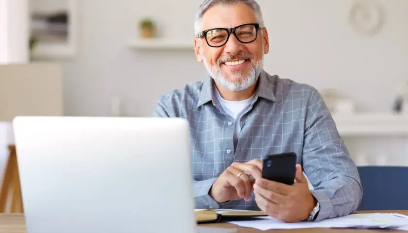 Uśmiechnięty senior z okularami pracuje przy laptopie i telefonie. To ulga dla pracujących seniorów, którzy cenią sobie nowoczesne rozwiązania.