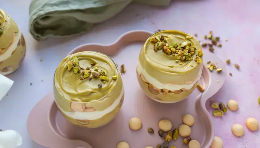 Dwa pucharki z deserem: krem pistacjowy z mascarpone, przełożony biszkoptami i bitą śmietaną, posypany pistacjami.