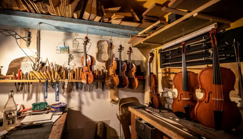 Laboratorio liutaio: violini, viole, violoncelli e archi appesi. Legname e attrezzi da lavoro, con un vasetto di kerria lacca.