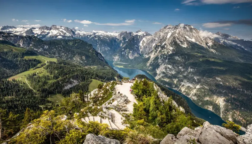 Atemberaubender Blick über den Königssee und die Berchtesgadener Alpen. Ein Aussichtspunkt mit Menschen lädt zum Verweilen ein.