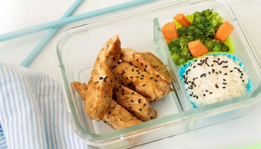 A delicious chicken bento box with teriyaki chicken strips, white rice sprinkled with black sesame seeds, and steamed broccoli with carrots.