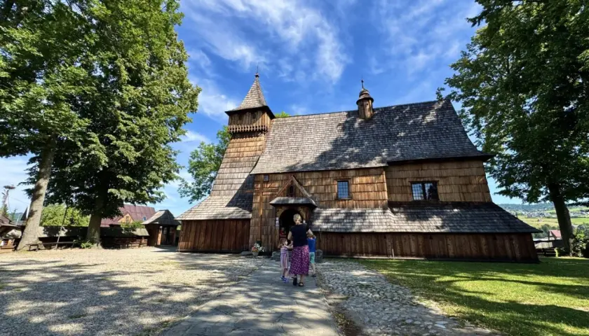 Drewniany kościół w Dębnie, z charakterystyczną wieżą i gontowym dachem, otoczony zielenią.