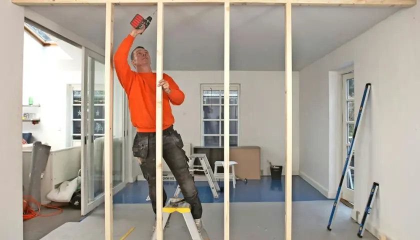 Man i orange tröja monterar träreglar för en ny vägg med skruvdragare.