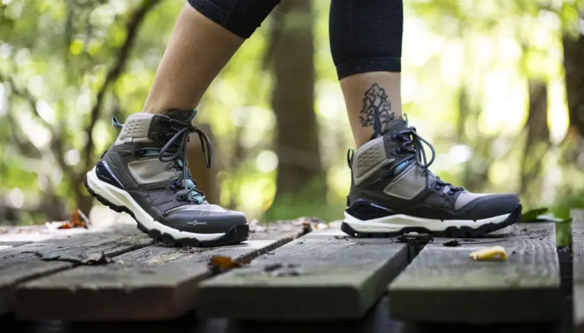 A person walks on a wooden path in the forest, wearing hiking boots. One foot half size bigger than the other, with a tree tattoo on the ankle.