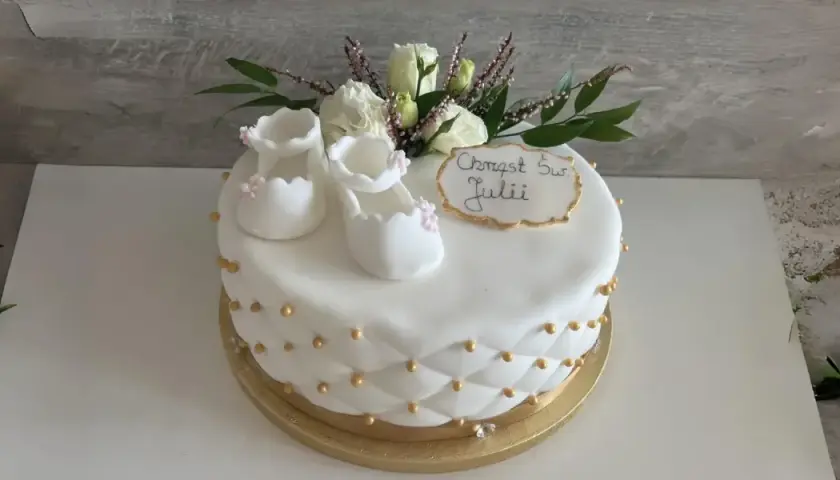 Delikatny tort na chrzest dla Julii, ozdobiony białymi bucikami, kwiatami i złotymi perełkami.