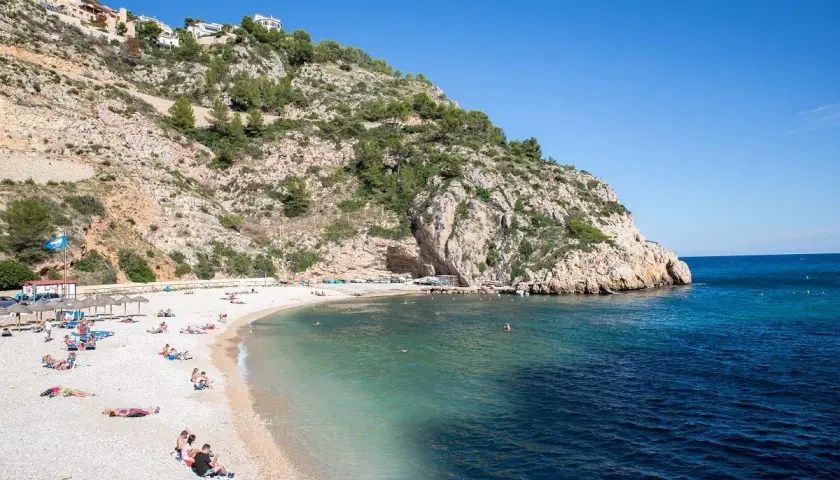 Gente disfrutando del sol en una playa de guijarros en la **Costa Blanca**, con aguas turquesas y un acantilado rocoso.