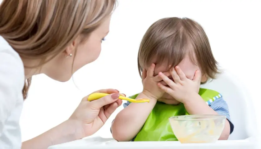 Mama próbuje nakarmić malucha, ale niemowlę nie chce jeść, zakrywając buzię rączkami.